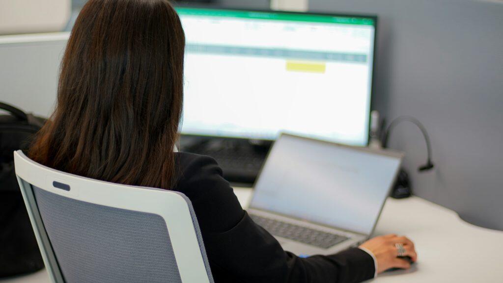 A professional workspace features a person seated at a desk, facing away from the camera. The individual, wearing a dark jacket, is focused on a laptop with a large monitor behind it displaying an application with a green header, possibly a spreadsheet. The scene hints at what spreadsheets to keep for a small business, showcasing an efficient setup. The desk is neat, with a black bag on the left and a dark cable on the right, suggesting an office environment focused on productivity.