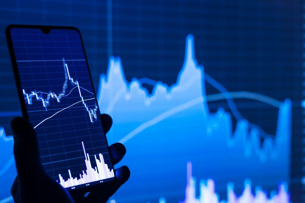 A close-up of a smartphone held in a silhouetted hand, displaying a detailed financial chart with candlestick patterns, trend lines, and volume indicators on a blue grid background. The phone screen is brightly lit, showing time markers like 03:00, 06:00, and 09:00 along the bottom axis. In the blurred background, a larger screen displays a simplified blue line graph, reinforcing the theme of financial data analysis for B2B LinkedIn product users. The scene is illuminated by the glow of the screens, creating a focused and tech-driven atmosphere.