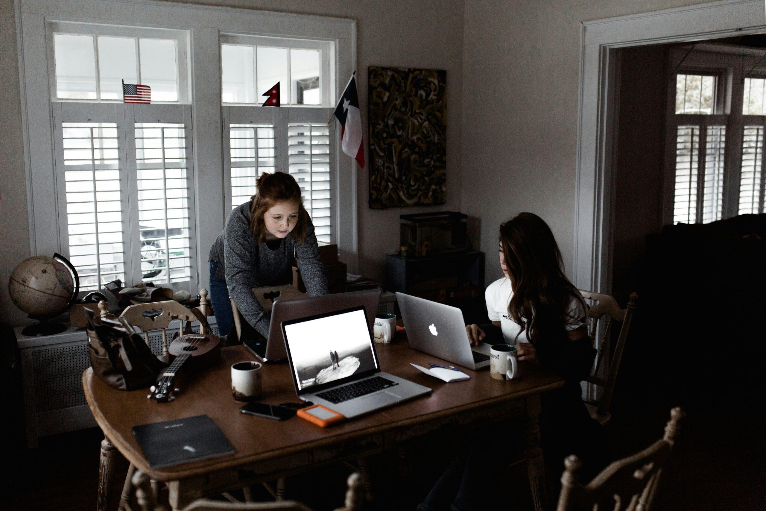 Two individuals working in a softly lit home office. A wooden table in the center holds two laptops (one with an Apple logo), a smartphone, an orange hard drive, a globe, a ukulele, two mugs, and notepads. Large windows with white shutters and a Texas flag on the wall add to the room's casual and collaborative ambiance. Flags and a globe hint at international themes, inspiring thoughts about the best small business to start.
