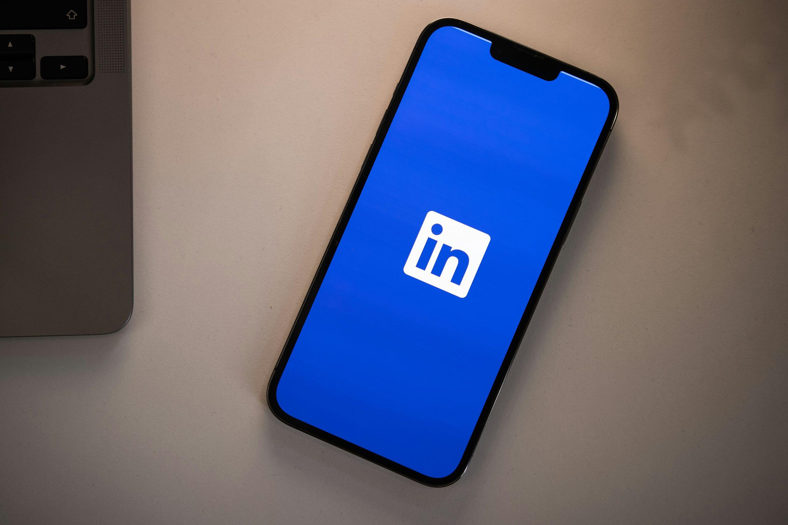 A top-down view of a smartphone displaying the LinkedIn logo on its bright blue screen, placed diagonally on a light-colored textured surface. A portion of a laptop, likely a MacBook, is visible in the upper-left corner, showcasing part of its keyboard and speaker grille. The composition is clean and modern, emphasizing technology in a B2B LinkedIn product professional workspace.