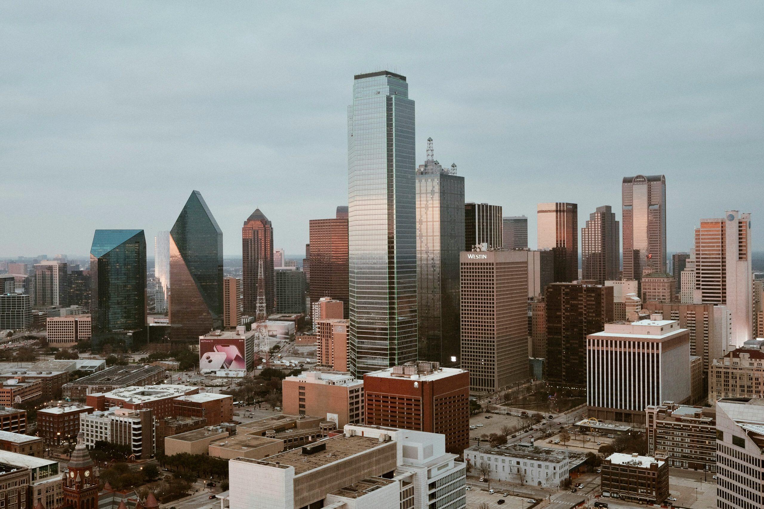 An aerial view of the Dallas, Texas skyline under an overcast sky, featuring a mix of modern skyscrapers and historic architecture. Prominent landmarks include the Bank of America Plaza with its green-lit outline, the blue-faceted Fountain Place, and the Museum Tower with its pyramid top. The Old Red Museum, a red brick building with a clock tower, is visible in the foreground. Corporate logos such as AT&T, Comerica, and Westin are displayed on buildings, while a billboard advertising the iPhone 13 is prominent at ground level. The scene captures the city's vibrant commercial and architectural diversity, making it a prime location for networking events Dallas.