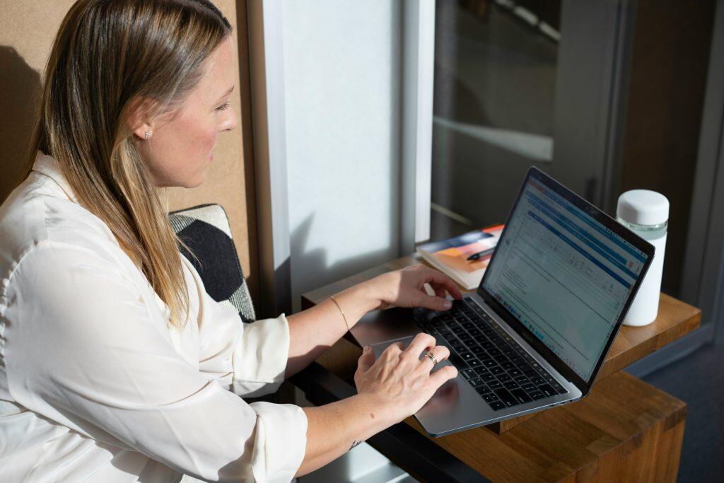 A side-profile view of a woman seated and working on a laptop in a bright, modern indoor setting. She has light brown hair with blonde highlights and is wearing a light-colored, collared shirt. The laptop, resembling a MacBook, is placed on a warm-toned wooden desk, displaying an email client on its screen. A white water bottle and an open notebook with a pen rest on the desk. The background features a mix of light and shadow, suggesting a professional yet comfortable workspace, perfectly suited for engaging with a B2B LinkedIn product.