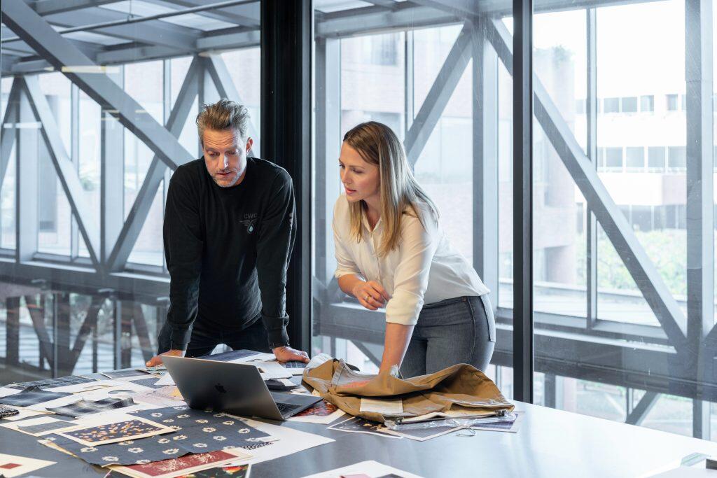 Two professionals, a man and a woman, collaborate over a large table covered with fabric swatches, papers, and a silver laptop. The man, wearing a black shirt with a 'CWC' logo, leans over the table, while the woman, in a white collared shirt, examines a khaki garment. The background features expansive windows with a modern architectural framework, offering a view of buildings and greenery outside. The scene conveys a focused and creative workspace, ideal for showcasing a B2B LinkedIn product.