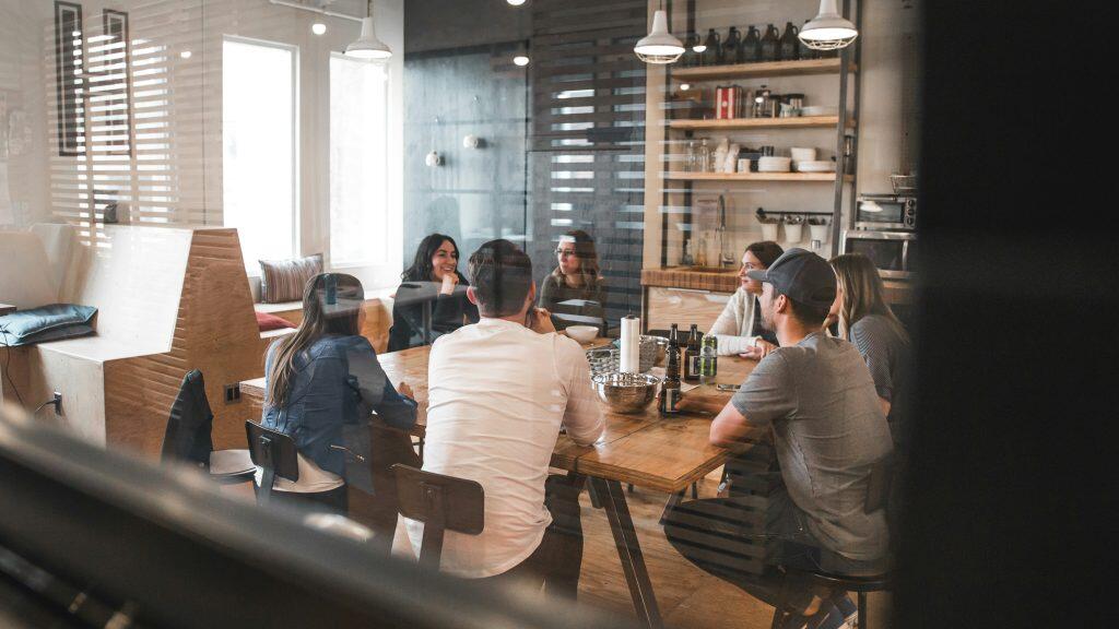 An indoor scene featuring a group of seven individuals gathered around a large wooden table in a modern communal space. The setting includes a mix of natural light from tall windows with horizontal blinds and artificial lighting from pendant fixtures. The table is scattered with items like beer bottles, a silver mixing bowl, and a roll of paper towels. In the background, a kitchenette with open wooden shelves displays plates, glassware, and large bottles, while a built-in bench with cushions lines the left wall. The image is viewed through a reflective glass partition, adding horizontal reflections that overlay the scene, creating a layered and dynamic visual effect. This setting is perfect for networking events Dallas, allowing attendees to connect in a stylish and open atmosphere.