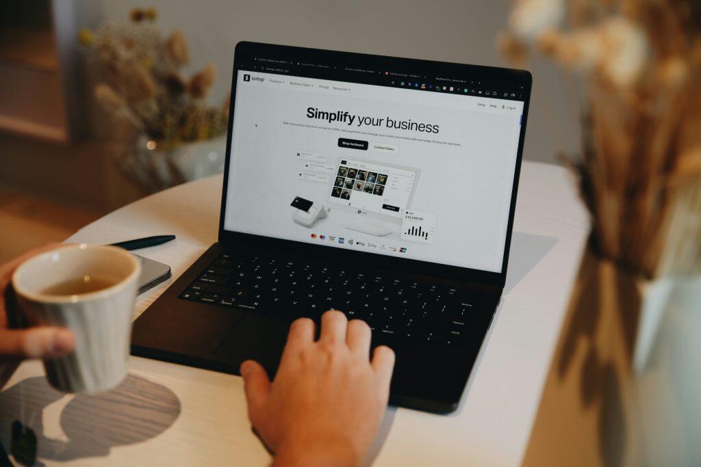 An overhead view of a person using a black laptop displaying the SumUp website, with the main heading 'Simplify your business' and calls to action like 'Shop hardware' and 'Contact Sales.' The laptop is on a light-colored table, with the user's right hand on the keyboard and their left hand holding a ribbed ceramic mug filled with a dark liquid. A smartphone and black pen are partially visible to the left of the laptop. The blurred background includes warm tones and decorative elements like dried flowers, creating a cozy and productive work environment, reflecting design trends for 2025 in web design and graphic design.
