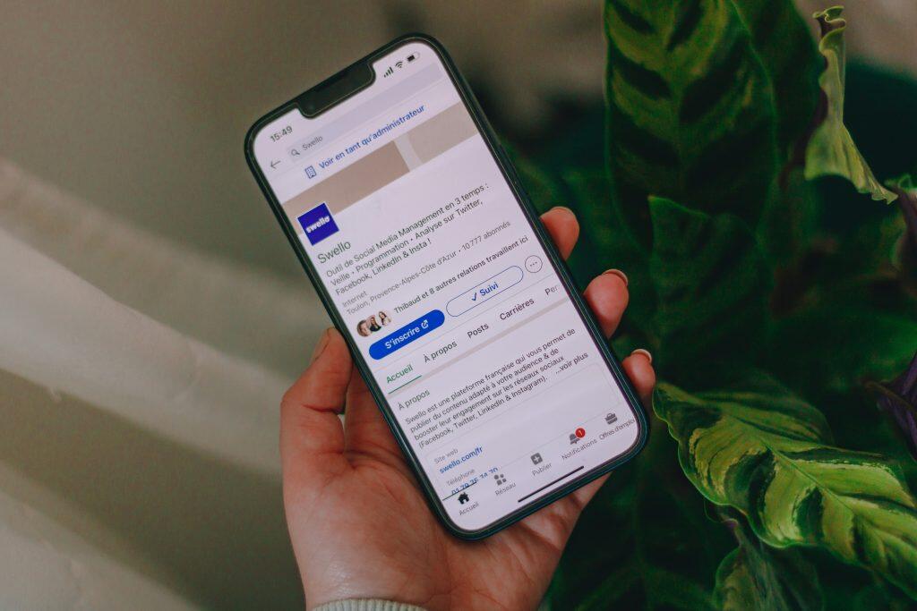 A close-up view of a smartphone held vertically in a person's left hand, displaying the profile page of 'Swello,' a French social media management platform. The screen shows details like the company logo, follower count, and navigation tabs, with a clean white and blue interface. The background features blurred green plant leaves and a light-colored surface, creating a professional yet natural aesthetic for a B2B LinkedIn product.