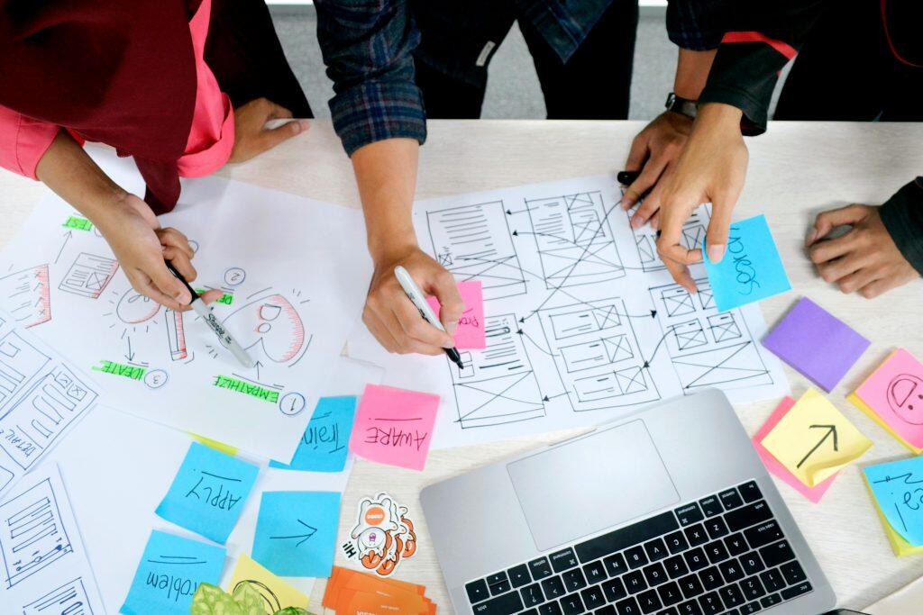 An overhead view of a collaborative UX design session with three individuals working on a light-colored table. Two large white sheets of paper display hand-drawn diagrams, along with wireframes and user flows. Colorful sticky notes with words like 'APPLY,' 'AWARE,' and 'Search' are scattered across the table. A silver laptop with a visible keyboard is positioned on the lower right, surrounded by stickers, orange business cards, and a small green plant. The participants’ hands are actively engaged, holding markers and sticky notes, creating a dynamic and creative workspace that embodies design trends for 2025 in web design and graphic design.