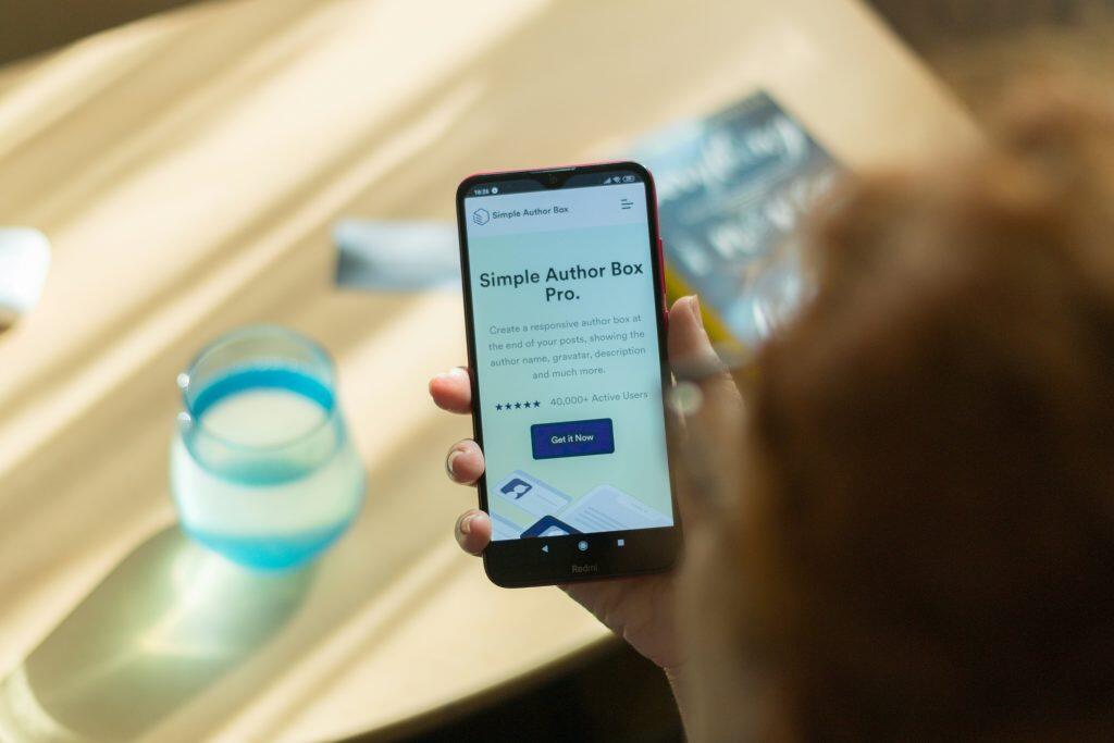A close-up of a red Redmi smartphone held vertically in a person's right hand, displaying a webpage for 'Simple Author Box Pro.' The screen features a heading, 'Create a responsive author box at the end of your posts,' along with a five-star rating, '40,000+ Active Users,' and a call-to-action button reading 'Get it Now.' The background is blurred, showing diagonal light and shadow patterns on a surface, with a glass containing a light blue liquid visible to the left. The setting conveys a casual and relaxed atmosphere, reflecting design trends for 2025 in web design and graphic design.