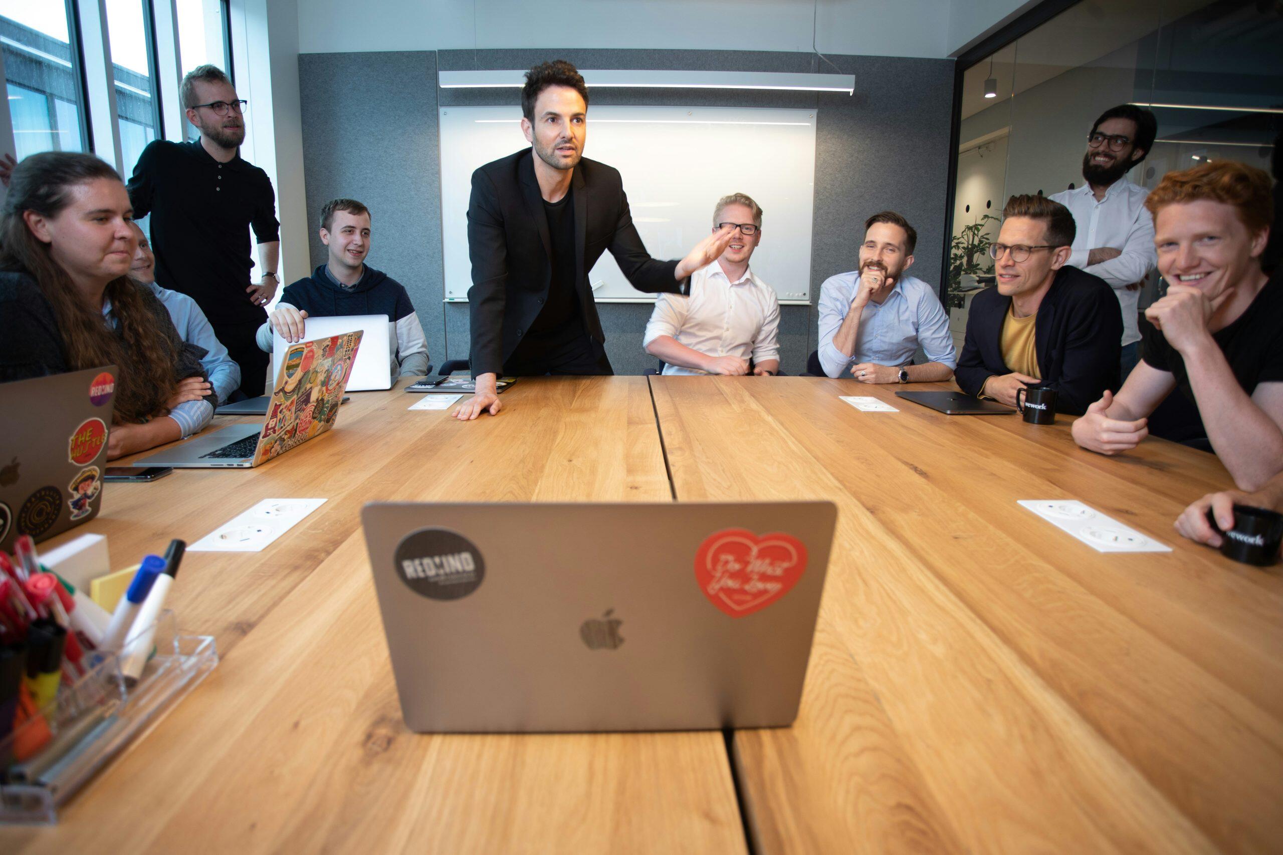 A group of nine people in a modern office, gathered around a wooden table with laptops, mugs, and office supplies, engaged in a collaborative business management meeting under natural light from large windows.