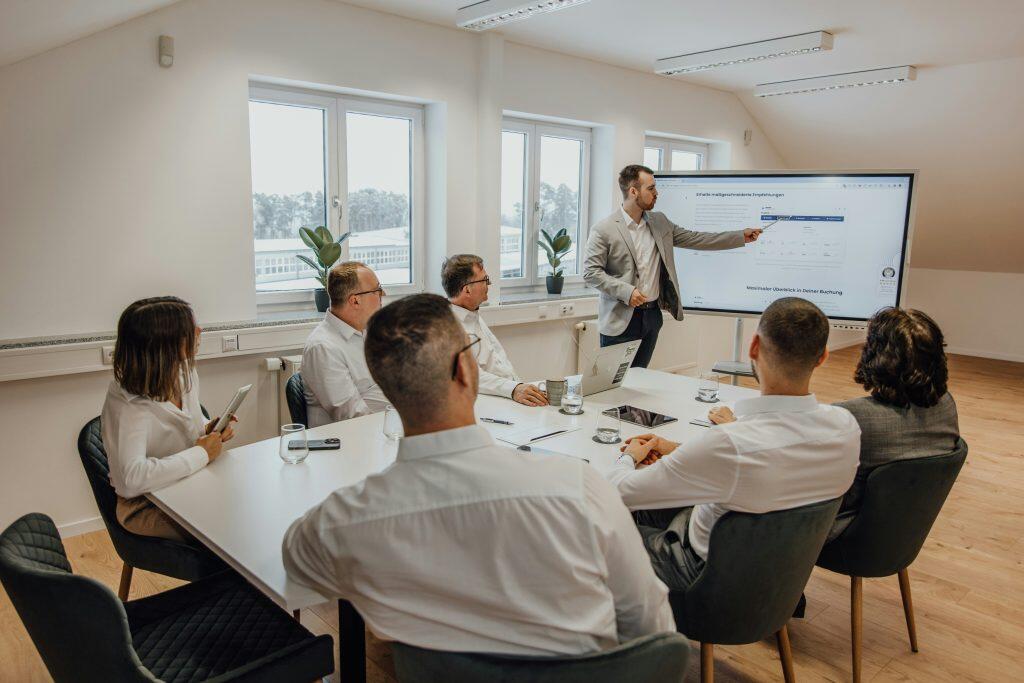 A professional meeting with seven people, one standing and presenting data on a digital screen, while six others sit around a white conference table with laptops, notebooks, and water glasses, in a bright room with large windows and potted plants, discussing business management strategies.