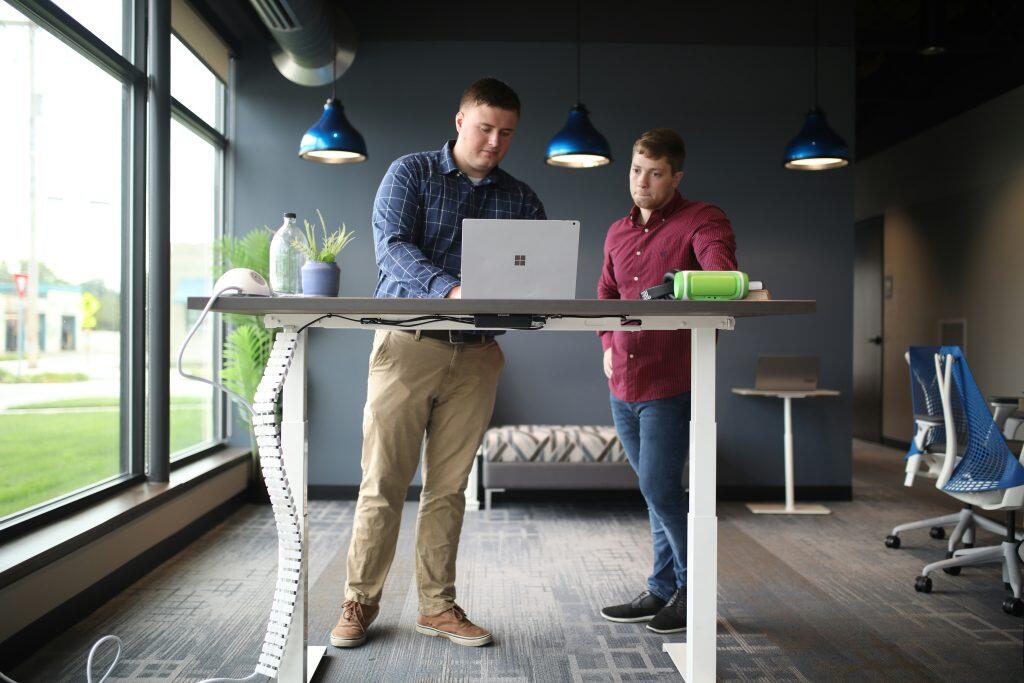 Two people standing at a modern height-adjustable desk, collaborating on a laptop, with a small plant and gadgets on the desk, in a well-lit office featuring pendant lights, a large window, and contemporary furniture, discussing business management.