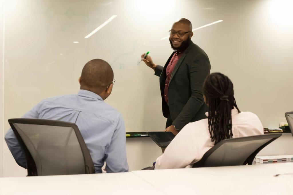 A professional setting with one person standing and presenting at a whiteboard using a green marker, while two others sit in office chairs facing the presenter, in a brightly lit business management office or classroom environment.