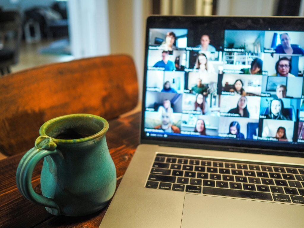A cozy remote work setup featuring a laptop on a wooden table, displaying a video call interface with multiple participants in a grid layout. To the left of the laptop, a blue-green ceramic mug adds a personal touch. The scene reflects a casual and comfortable environment, ideal for online meetings, virtual collaboration, or networking events Las Vegas.