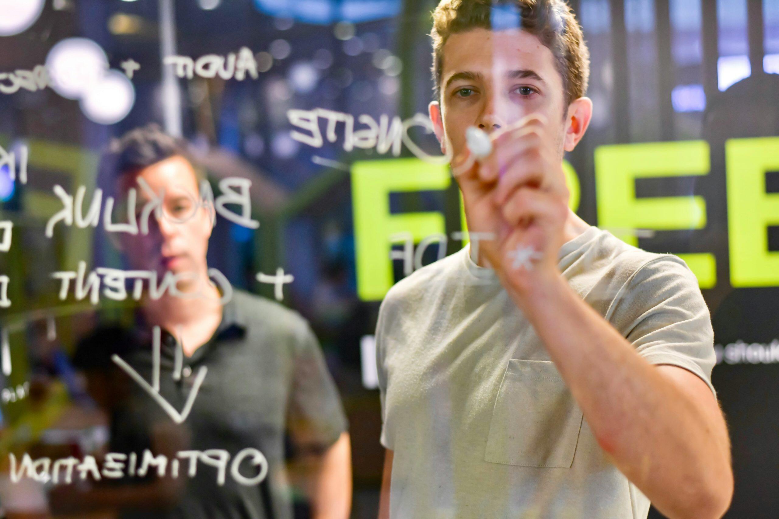 Two individuals in a professional setting, one writing on a transparent surface with a white marker while the other observes. The surface displays handwritten terms like 'AUDIT,' 'CONTENT,' 'STRATEGY,' 'BACKLINK,' 'OPTIMIZATION,' and 'DIRECT RESPONSE MARKETING,' with some symbols like plus signs and arrows. In the blurred background, the word 'FREE' is prominently visible in bold green letters, suggesting a brainstorming or planning session focused on digital marketing, DIRECT RESPONSE MARKETING, or SEO strategies.