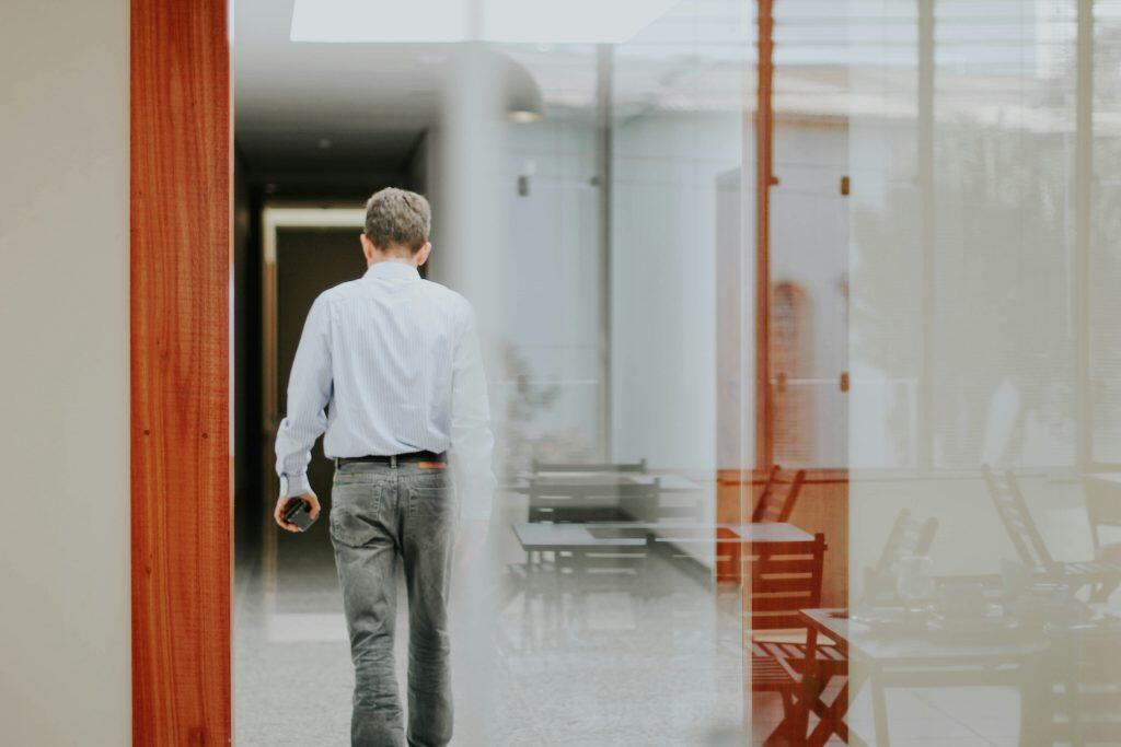 A man in a light blue shirt and grey trousers walking down a modern indoor corridor with reflective glass surfaces and visible furniture, suggesting an office or professional setting, possibly heading to discuss exit interview questions.