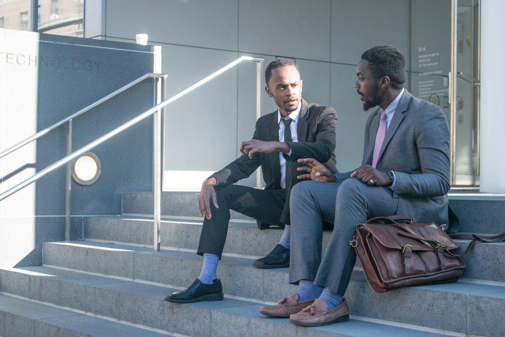 Two men in business attire seated on concrete steps outside a building, with a brown leather bag, and coffee cup, discussing conferences in Las Vegas this week.