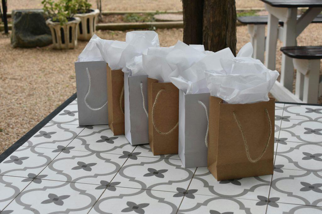 An outdoor scene featuring six gift bags, perfect for trade show giveaways, arranged in a row on a decorative tiled table. The bags alternate in color between light gray and brown kraft paper, each filled with crumpled white tissue paper. The table has a quatrefoil tile pattern in light gray and white with dark gray accents. The background is softly blurred, showing a tree trunk, gravel ground, decorative planters with greenery, and parts of outdoor picnic tables or benches.