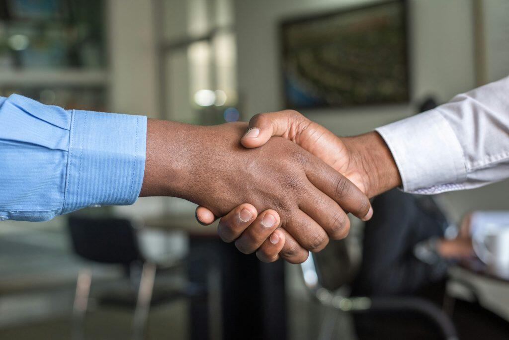 A close-up image of two individuals shaking hands, symbolizing agreement or partnership. The left hand, wearing a light blue striped dress shirt, is on top, while the right hand, wearing a white dress shirt, is underneath. The background is softly blurred, suggesting an indoor office setting with hints of furniture, ambient lighting, and muted tones of gray, green, and black. The focus is on the clasped hands and forearms, emphasizing the gesture of connection, often seen at trade show giveaways.
