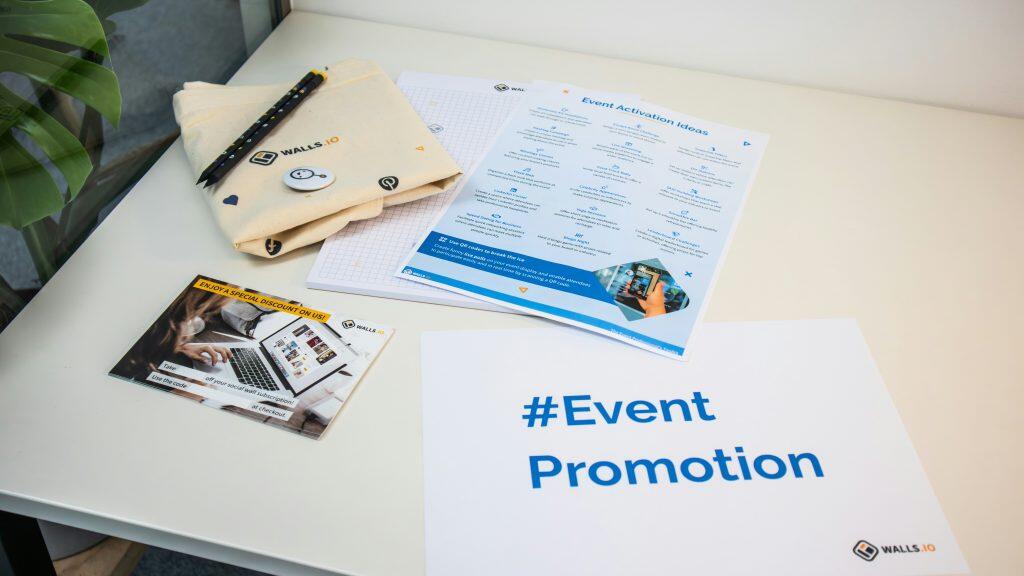 A tabletop scene showcasing promotional materials from 'Walls.io,' including a beige canvas bag with 'WALLS.IO' printed on it, two black pencils, and a button. Nearby, a blue and white 'Event Activation Ideas' sheet lists creative event engagement ideas with icons. A discount flyer offers a special deal on a social wall subscription, and a large white sheet displays '#Event Promotion' in bold blue text. The trade show giveaways are neatly arranged on a white table, with a green Monstera plant partially visible in the corner, suggesting a professional and organized event marketing setup.
