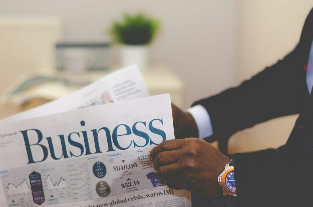 A businessman in a dark suit holds a financial newspaper with the headline "Business" prominently displayed. The newspaper features sections on currencies, commodities, and market trends, including exchange rates, gold prices, and stock data. The man's wristwatch, with an orange bezel and intricate design, is visible. The background is blurred, showing a green potted plant and a light-colored wall, emphasizing the focus on the newspaper and raising the question how much do brokers charge to sell a business.