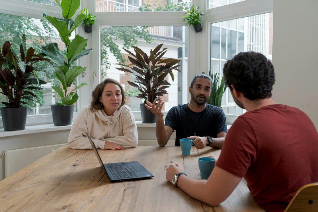 A candid indoor scene featuring three individuals seated around a large wooden table in a bright, plant-filled room. A woman on the left, wearing a beige hoodie, is seated with a black laptop in front of her. A man in the middle, with dark skin and a beard, gestures while holding a light blue mug. Another man, seen from behind, listens attentively. The background includes a large bay window with white frames, potted plants, and outdoor greenery visible through the glass. The setting suggests a casual meeting or collaborative work session, perhaps brainstorming ways to find angel investor Los Angeles.