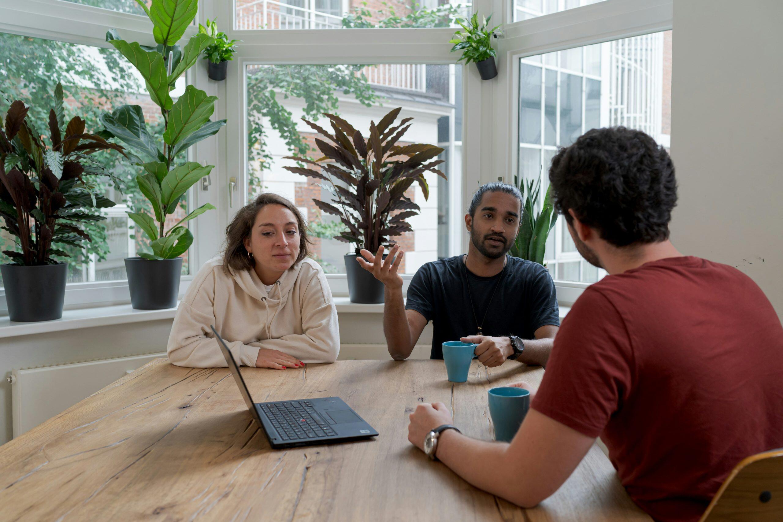 A candid indoor scene featuring three individuals seated around a large wooden table in a bright, plant-filled room. A woman on the left, wearing a beige hoodie, is seated with a black laptop in front of her. A man in the middle, with dark skin and a beard, gestures while holding a light blue mug. Another man, seen from behind, listens attentively. The background includes a large bay window with white frames, potted plants, and outdoor greenery visible through the glass. The setting suggests a casual meeting or collaborative work session, perhaps brainstorming ways to find angel investor Los Angeles.