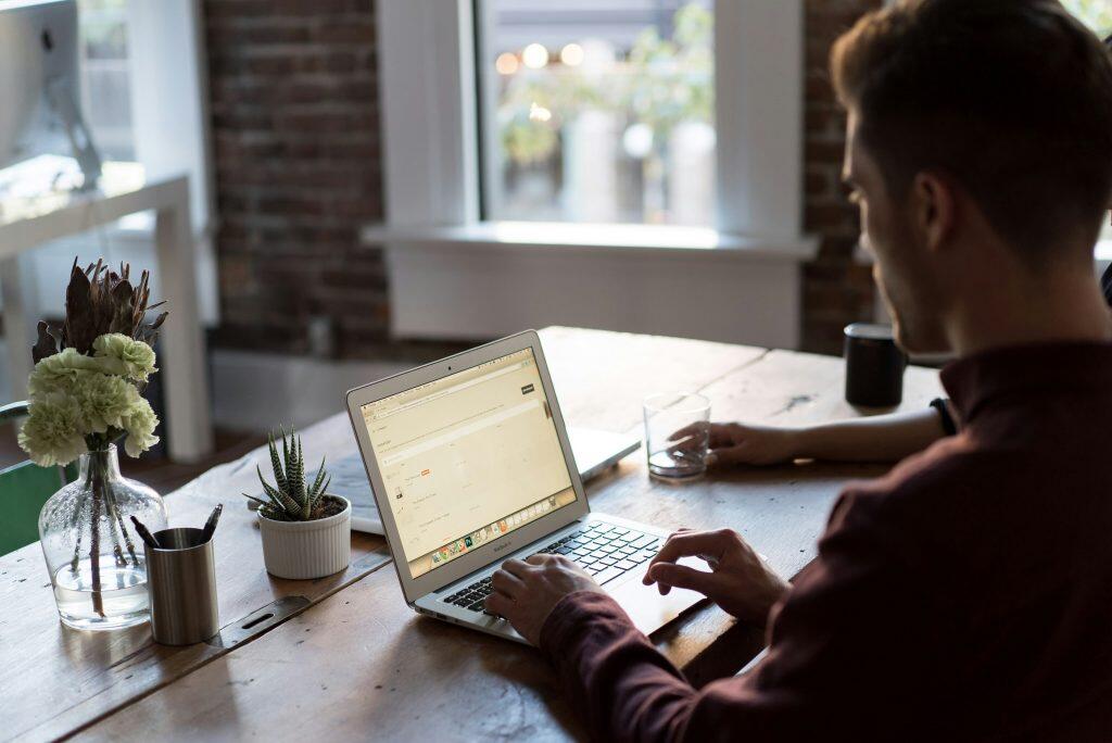An over-the-shoulder view of a person working on a silver MacBook Air laptop at a rustic wooden table. The laptop screen displays an e-commerce inventory management interface, perhaps tracking investments to find angel investor Los Angeles. The person, wearing a reddish-brown long-sleeved shirt, has their hands on the keyboard and trackpad. The table is adorned with a clear vase of green flowers, a pen holder, a small succulent plant in a white pot, and a second person’s hand holding a glass of water in the background. Bright natural light streams through a large white-framed window, with blurred greenery and a brick wall visible in the background, creating a warm and productive atmosphere.