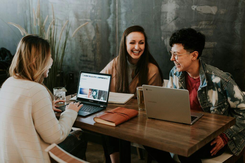 Three young adults are seated around a wooden table in a modern, casual setting, laughing and enjoying a lively moment together, perhaps at one of the lively networking events Philadelphia. The table holds laptops, notebooks, and drinks, suggesting a collaborative or social activity. One person wears a ribbed cream top, another a beige jacket, and the third a tie-dye denim jacket with glasses. The background features a textured wall and decorative plants, adding a cozy, contemporary vibe.
