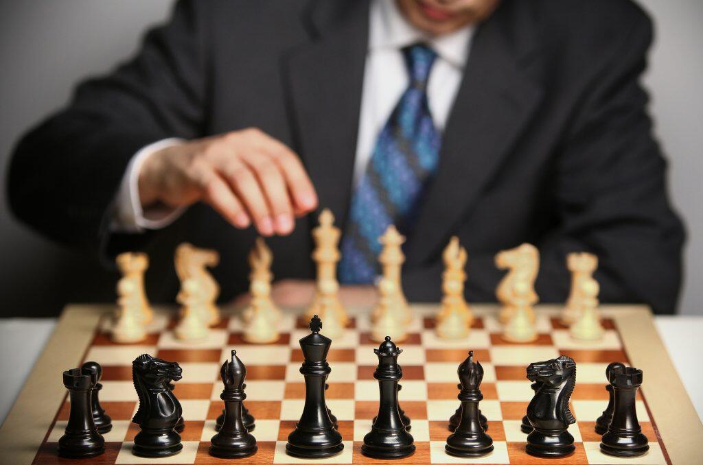 A chessboard with black pieces in the foreground and white pieces in the background, a person in formal attire reaching towards the board, suggesting a move in a strategic game, as though seeking AI answers for the next move.