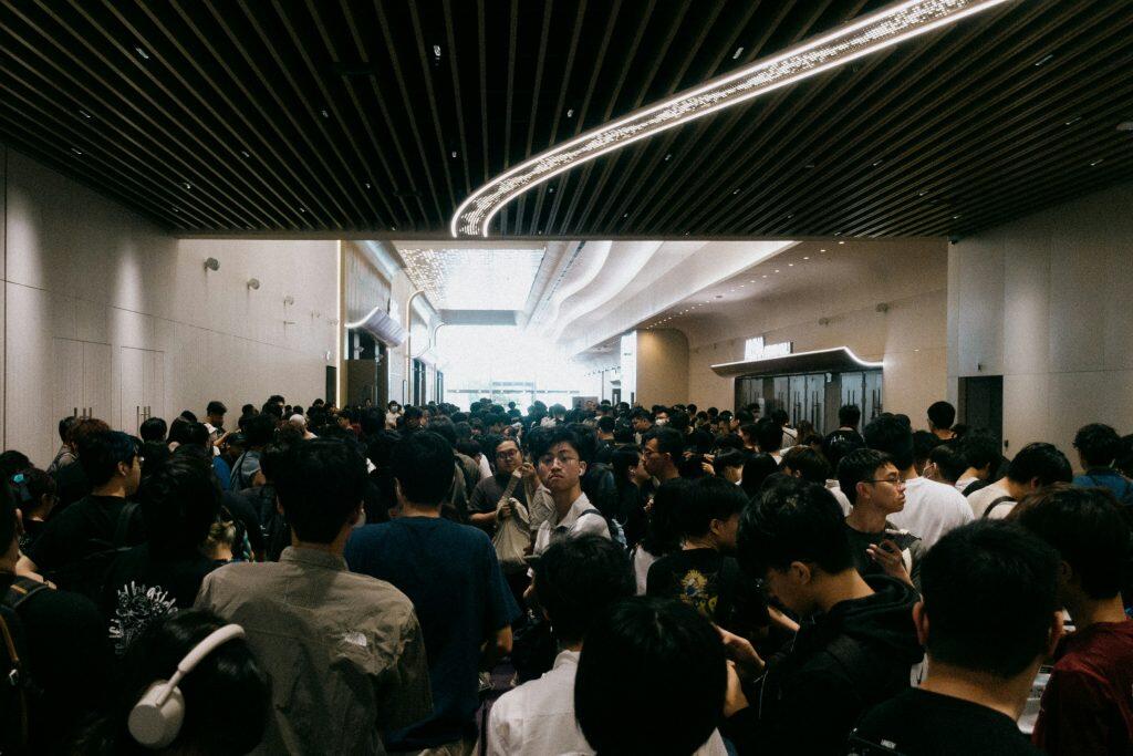 A crowded hallway filled with people, many wearing casual attire, backpacks, or headphones, moving or waiting. The modern space features a ceiling with parallel wooden slats and a curved illuminated strip, providing soft lighting. Plain light-colored walls with small, evenly spaced lights line the hallway. Glass doors at the far end allow natural light to brighten the area, suggesting an exit or transition to another space. The scene conveys a contemporary, bustling environment, possibly at events in Phoenix or a public venue.