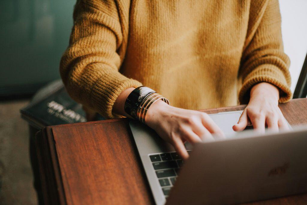 Close-up of a person typing on a silver laptop placed on a dark wooden table. The individual is wearing a chunky mustard yellow knit sweater, with metallic bracelets adorning their left wrist. The background is softly blurred, featuring a muted teal wall and hints of light, creating a cozy and focused atmosphere, perfect for researching networking events Philadelphia.