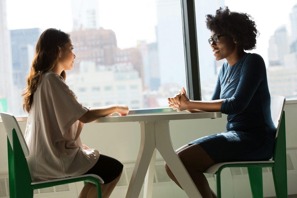 Two women sit across from each other at a modern white table in a brightly lit room with a large window overlooking a city skyline. The woman on the left, with long wavy brown hair, wears a light-colored top and dark pants, resting her arm on the table and looking attentively at the other woman. The woman on the right, with dark curly hair and glasses, wears a teal long-sleeved top and a heart-shaped necklace, smiling warmly with her hands clasped on the table. The Client Success Manager sits confidently, illustrating their attentive approach. The chairs have white seats with vibrant green metal frames, adding a pop of color to the neutral-toned space. The window in the background floods the room with natural light, showcasing a blurred cityscape and a bright sky.