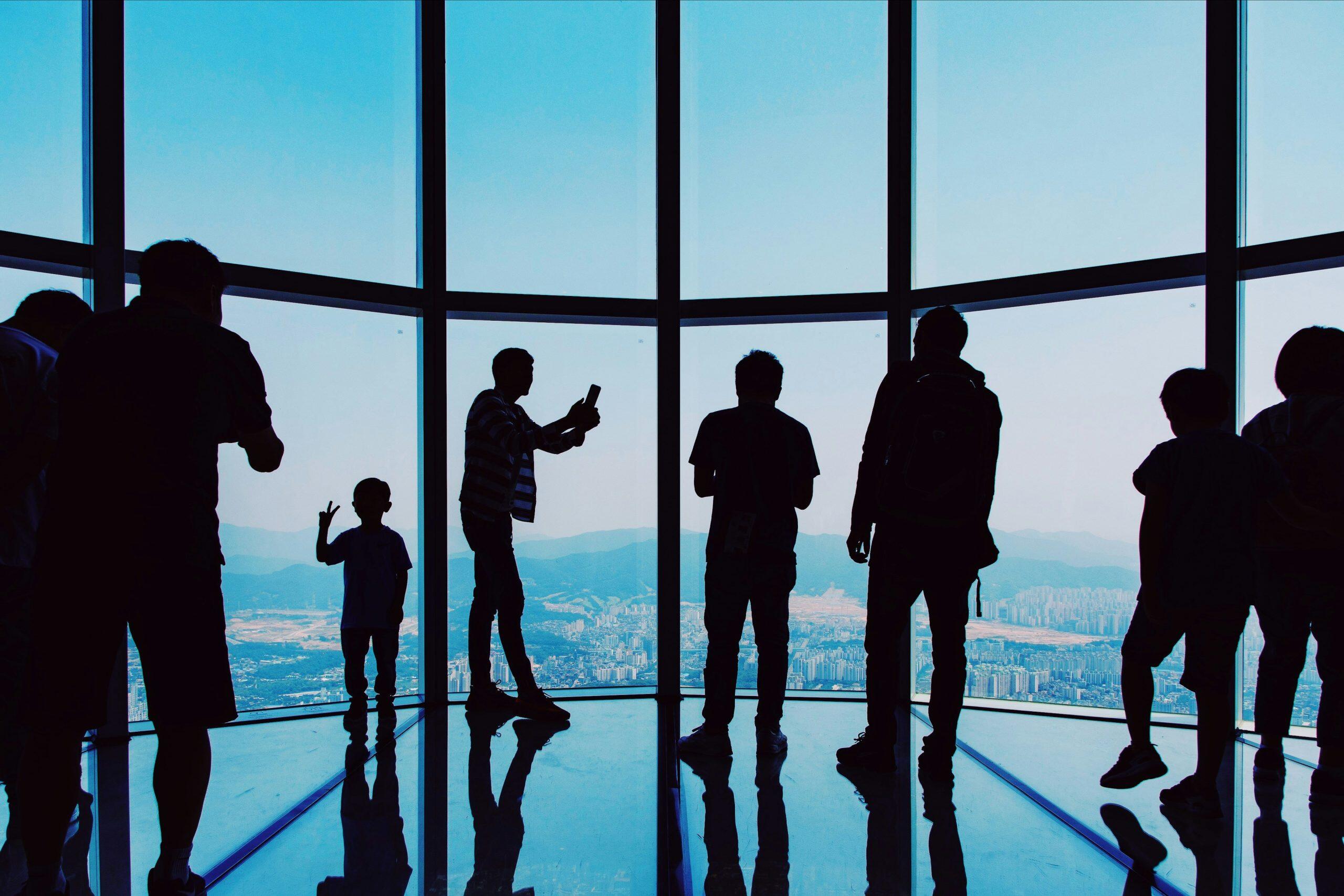 Silhouetted figures, including a child making a peace sign, stand in front of large curved windows in a high-rise building. The windows reveal a panoramic view of a cityscape with numerous buildings and mountains in the distance. The reflective floor mirrors the silhouettes, adding depth to the scene. The image has a blue hue, emphasizing the bright natural light and the contrast between the urban and natural elements, much like the backdrop for events in Phoenix.