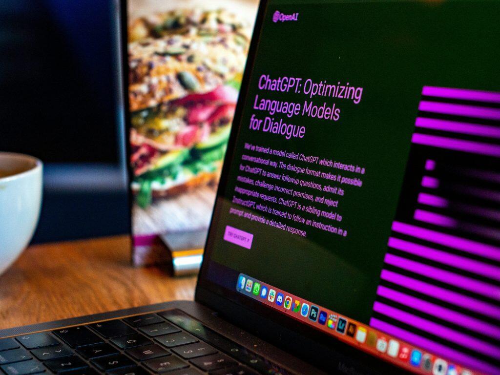 A laptop screen displaying information about ChatGPT by OpenAI, with text describing its conversational abilities and AI answers, set in a casual café-like environment with a blurred sandwich and a white cup in the background.