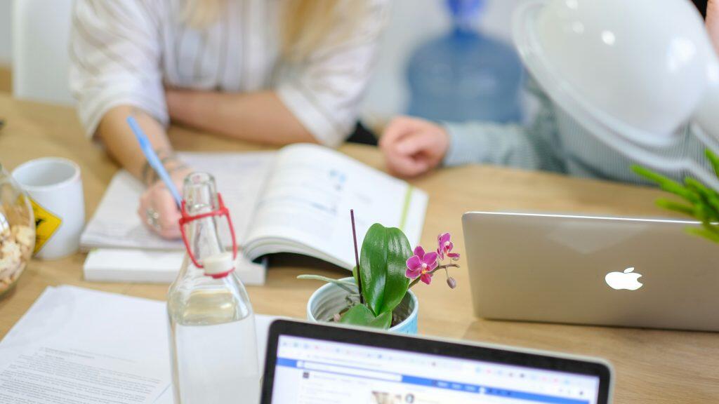 A modern workspace with a light wooden table featuring a white Apple laptop, a potted orchid with vibrant pink flowers, a glass water bottle with a red hinged cap, and a white mug with a yellow hazard symbol. A second laptop screen displays a web page, perhaps for an AI Agency, partially visible in the foreground. In the background, two individuals are blurred—one writing with a blue pen and another near a large blue water dispenser. The scene is brightly lit, blending digital tools and personal touches in a productive environment.