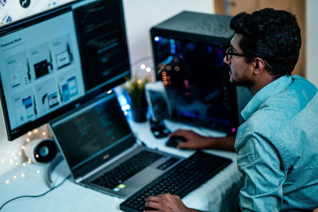 A workspace featuring a person seated at a desk with a dual-screen setup. The large monitor on the left displays a webpage with the heading 'Featured Work,' while the laptop in front of the person shows code. The individual, wearing glasses and a light blue shirt, is using a keyboard and mouse. The setup includes a desktop computer tower with RGB lighting, string lights for ambiance, a small speaker, and a potted plant. The scene suggests a tech-savvy and organized environment, focused on programming, best small business management software, or design work.