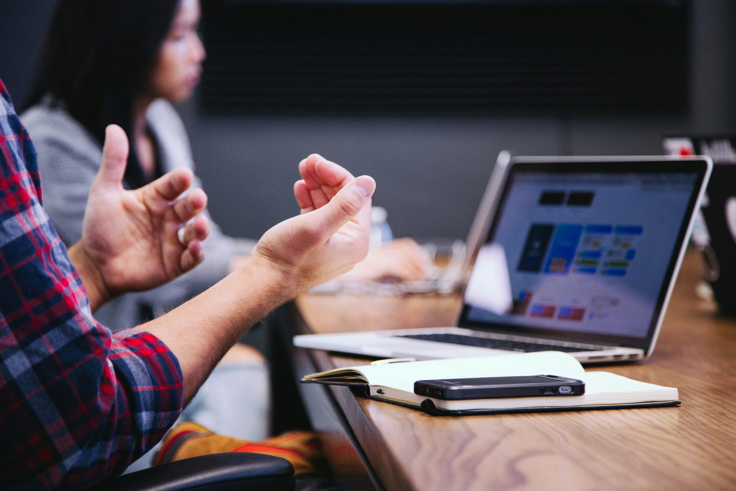 A person in a plaid shirt sits at a table, gesturing with their hands in a conversational manner. On the table, there is an open notebook with a smartphone placed on top, and a laptop with an unclear screen displaying a presentation or website. Another person is visible in the background, slightly out of focus, suggesting a group discussion or meeting in a casual, tech-savvy small business consulting setting.