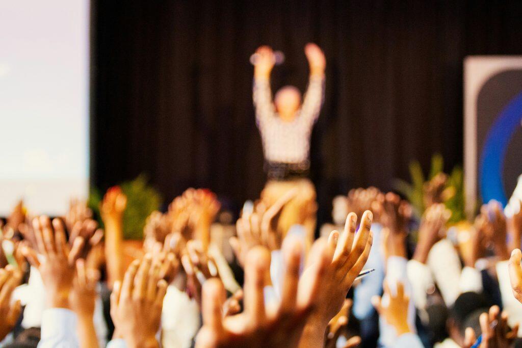 An interactive scene featuring numerous raised hands of varying skin tones in the foreground, symbolizing audience engagement. In the background, a blurred figure with arms raised stands centrally, likely a speaker or presenter. The setting includes dark curtains, a partially visible screen on the left, and green plants on either side, suggesting an indoor environment such as a seminar, workshop, or networking events Los Angeles. The composition emphasizes active participation and enthusiasm.