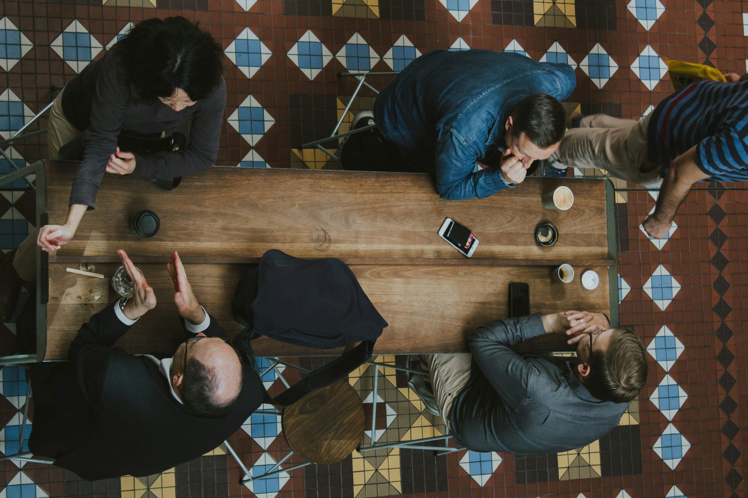 Overhead view of a casual meeting with four individuals seated around a rectangular wooden table and one person standing nearby. The table holds several cups, a smartphone, and a black bag. The floor features a colorful geometric tile pattern with red, blue, yellow, and white diamond shapes, adding visual interest to the setting—perfect inspiration for networking events phoenix.