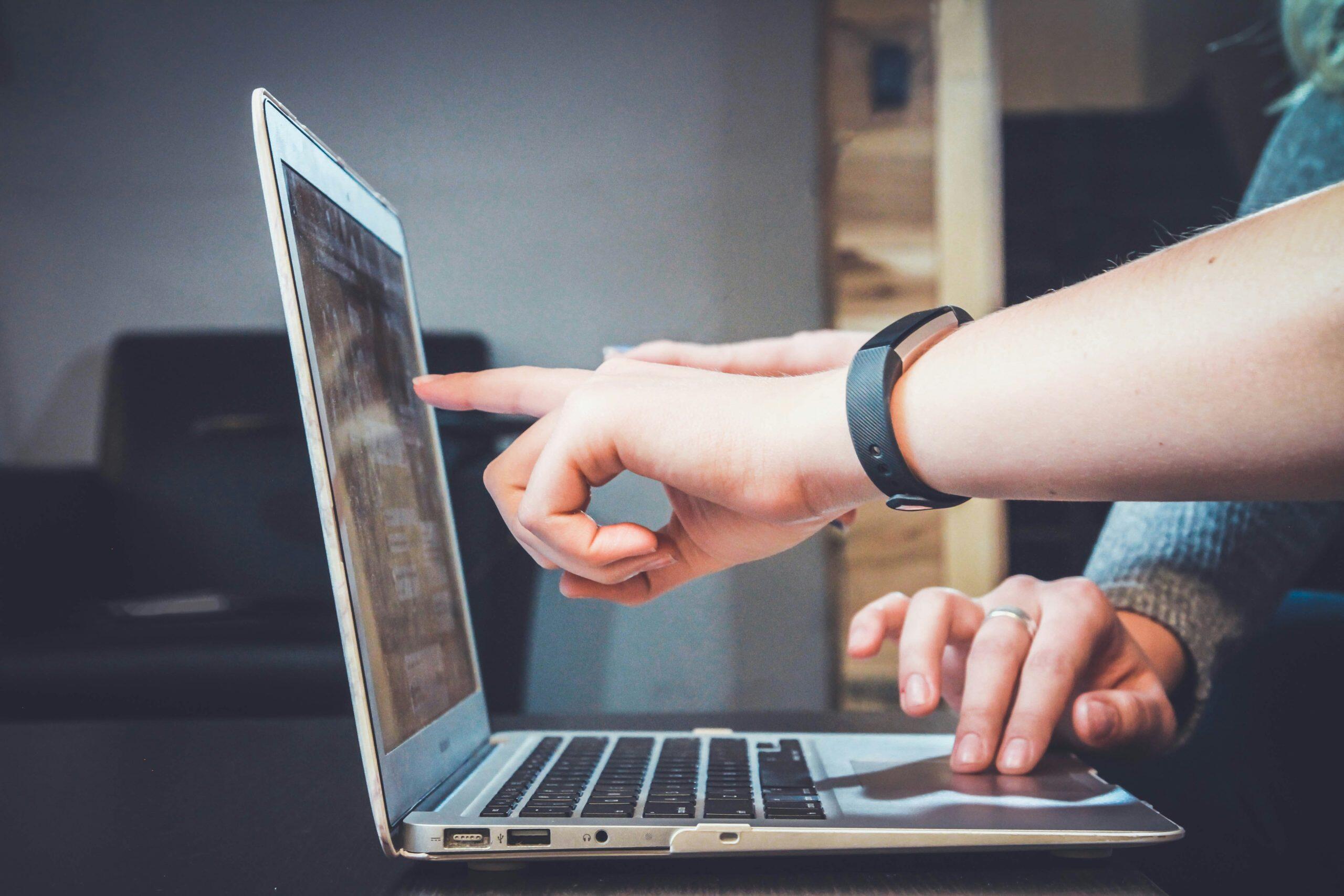 An indoor workspace scene featuring a laptop at the center. Two hands interact with the laptop—one pointing at the screen and the other positioned over the keyboard, suggesting collaboration or instruction. A black wristband, possibly a fitness tracker, is visible on the pointing hand. The background is blurred, emphasizing the interaction with the laptop and the best small business management software.