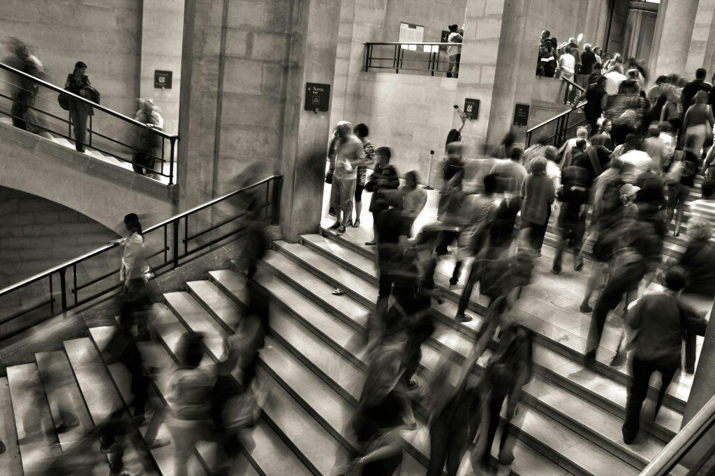 A bustling scene of people navigating a grand stone staircase in a public space. The staircase is wide, with metal railings on either side, and surrounded by tall, classical stone walls. Motion blur captures the movement of individuals dressed in casual attire, emphasizing the lively atmosphere. Signs reading 'Sortie' and 'Exit' are visible on the walls, indicating directions. The image is in black and white, adding a timeless and classic feel, reminiscent of the energy found at events in Phoenix.