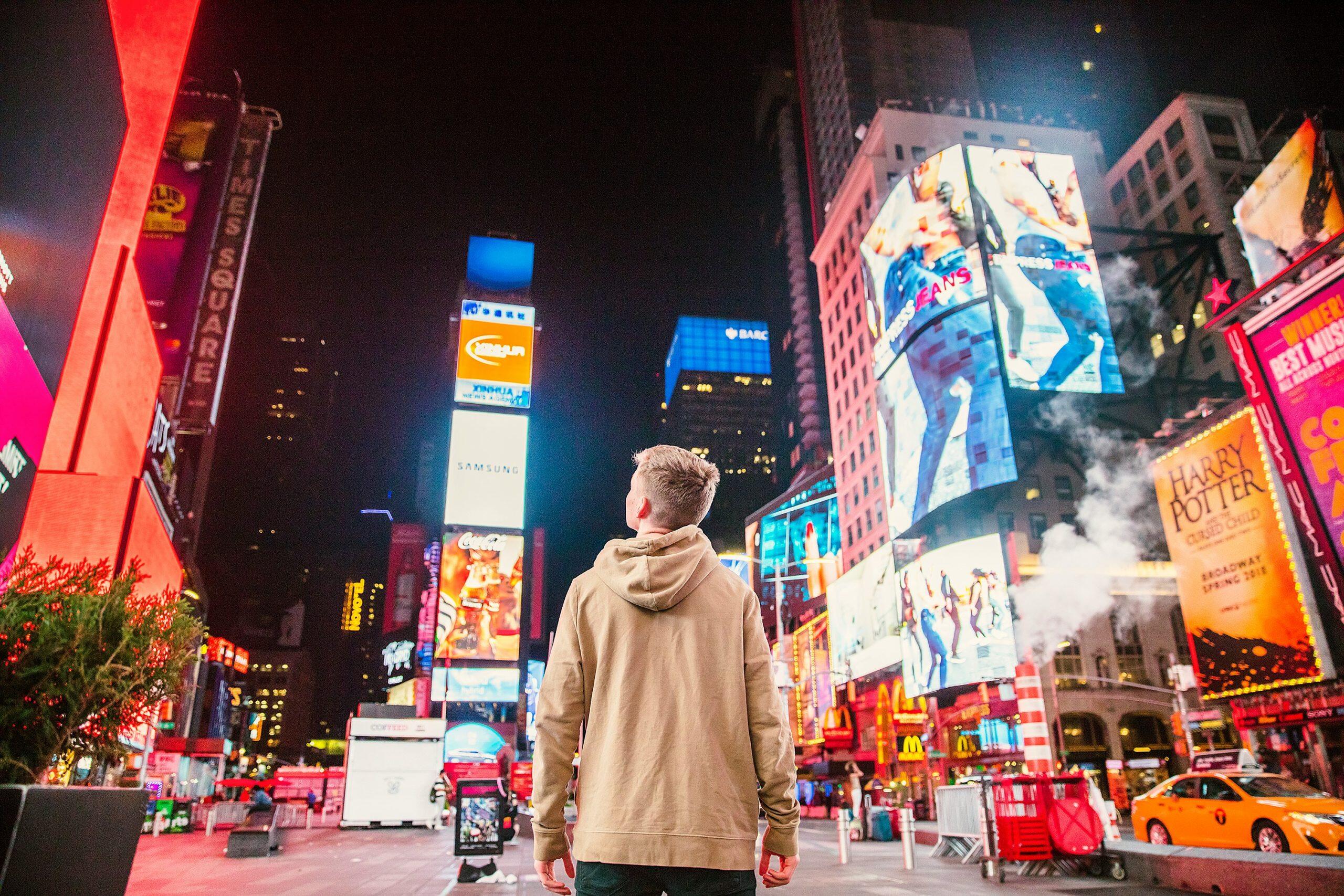 A person in a tan hoodie stands in Times Square, surrounded by vibrant billboards showcasing iconic brands and entertainment, symbolizing the best local advertising for small business in a bustling urban setting.