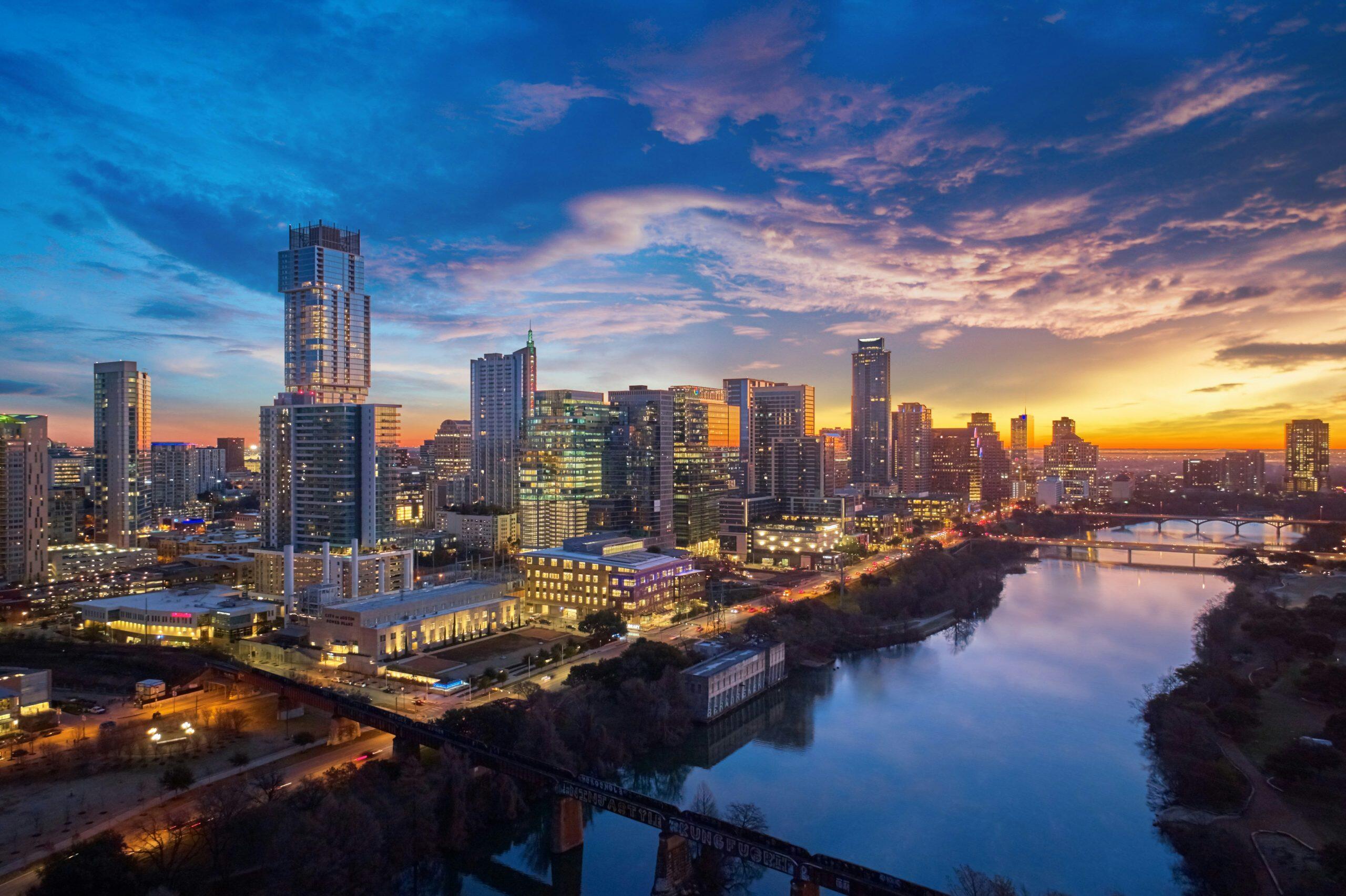 An urban cityscape at twilight featuring a skyline of modern skyscrapers silhouetted against a gradient sky transitioning from deep blue to orange and yellow. A river flows diagonally through the city, reflecting the sky's colors, with several bridges crossing it. This vibrant setting could inspire networking events in Austin, where professionals gather amidst a stunning backdrop to connect and collaborate. Bright city lights, including streetlights and car lights, create dynamic lines of light. Graffiti reading is visible on a bridge in the foreground, adding an urban artistic touch.