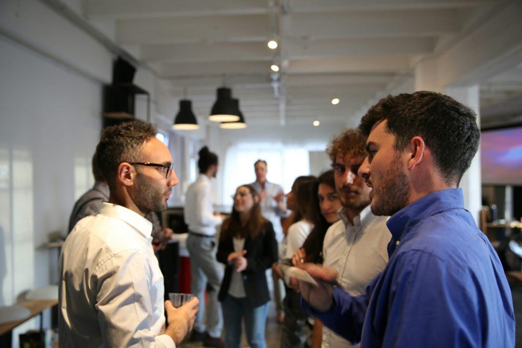 A casual indoor networking events Los Angeles or social gathering featuring a group of individuals engaged in conversation. Two men in the foreground are the focal point, one wearing a white shirt and glasses holding a clear cup, and the other in a blue shirt holding a phone. The background shows others mingling, some holding drinks, in a modern space with pendant lights, a large window with curtains, and visible furniture like chairs and tables. The warm lighting creates a welcoming and informal atmosphere.