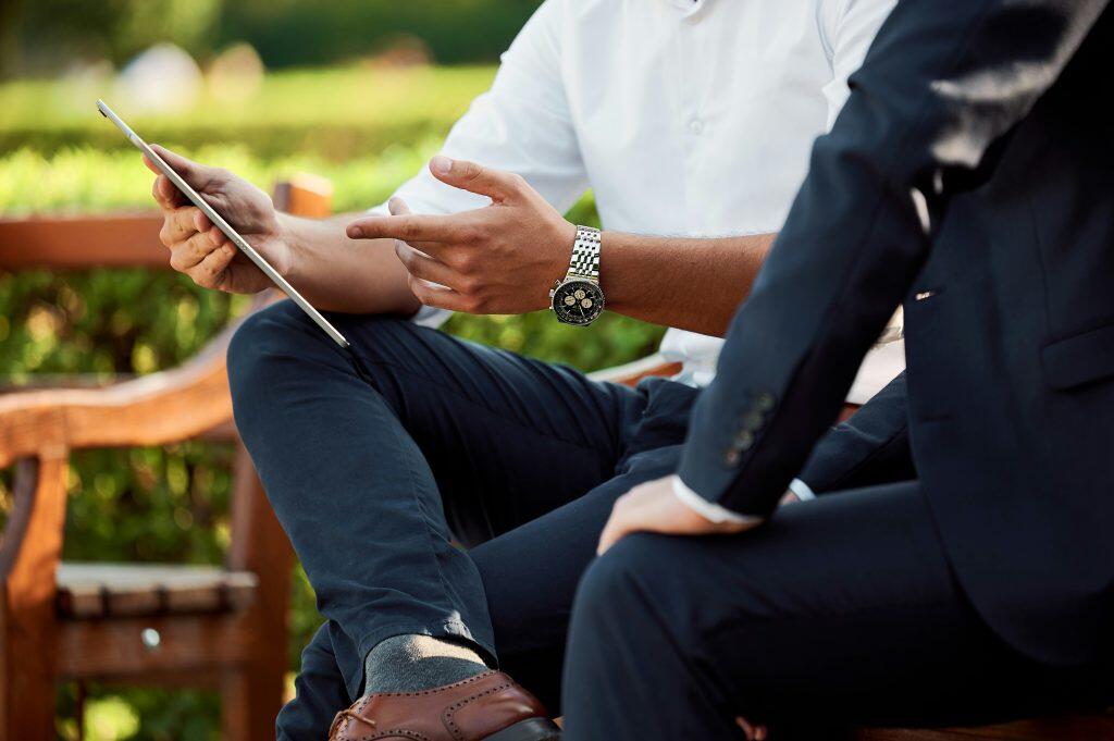 Two individuals are seated on a wooden bench in an outdoor setting with greenery in the background. The person on the left, wearing a white shirt, dark pants, and brown leather shoes, holds a tablet horizontally and gestures with their right hand. They are also wearing a silver watch with a dark face. The person on the right, dressed in a dark suit, appears to be engaged in the conversation. The scene suggests a professional or casual small business consulting meeting outdoors, with the tablet indicating a discussion involving digital content..