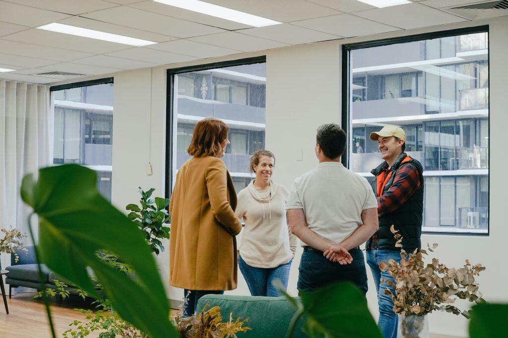 Four individuals are engaged in a casual conversation in a modern indoor setting. One person in a brown coat and another in a white shirt have their backs to the camera, while a person in a white sweater smiles at the group. A fourth individual, wearing a plaid shirt, vest, and cap, also smiles while facing the group. The space features large windows with views of an adjacent building, wooden flooring, and several green plants, creating a bright and welcoming atmosphere—perfect for networking events Los Angeles.