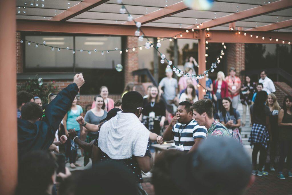 An outdoor social gathering under a pergola adorned with glowing string lights, creating a warm and festive atmosphere. The scene features a diverse group of people mingling and engaging in conversation. In the foreground, a person in a light gray hoodie draped over their shoulders faces away, while others gesture and interact. The midground highlights two men, one in a striped polo shirt with a lanyard and another in a colorful shirt, laughing together. The background shows a brick building with large windows and a group of smiling attendees. Floating bubbles add a playful touch to the lively and celebratory setting, perfect for networking events Atlanta.