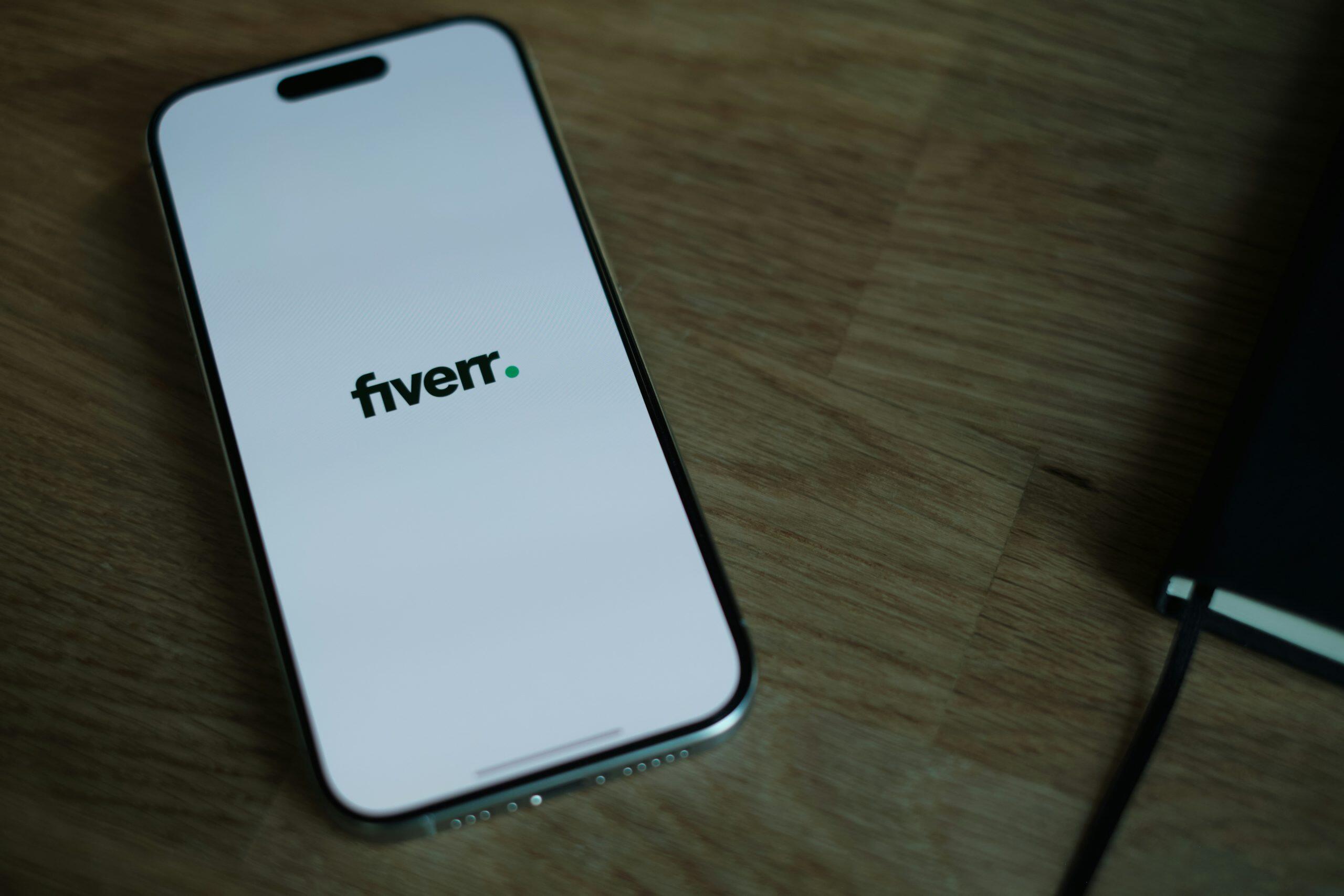 A smartphone placed diagonally on a wooden surface displays the Fiverr digital marketing logo prominently on its screen, with a small green dot following the text. To the right, part of a black notebook with a ribbon bookmark is partially visible, suggesting a professional workspace.