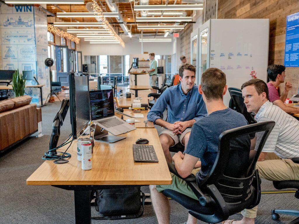 A modern open-plan office with wooden ceilings and contemporary lighting. In the foreground, three people sit at a desk with computer monitors displaying code, a laptop, and office supplies, engaged in discussion. Two more individuals are visible in the background at separate desks. The left wall features motivational text like 'WIN' and 'CUSTOMER SUCCESS,' while green plants and casual seating add a relaxed touch to the Fiverr digital marketing collaborative workspace.