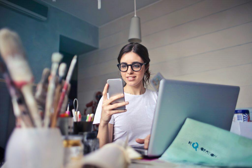 An image of a woman sitting at a desk in a creative workspace. She is wearing glasses and a white t-shirt, holding a smartphone in her right hand while her left hand rests on a laptop keyboard. The desk features blurred art supplies in the foreground, suggesting a creative environment. A light green cloth or paper partially covers the laptop. The background includes a wall with horizontal lines and a ceiling light fixture above, creating a cozy and modern atmosphere, perfect for exploring Fiverr digital marketing opportunities.